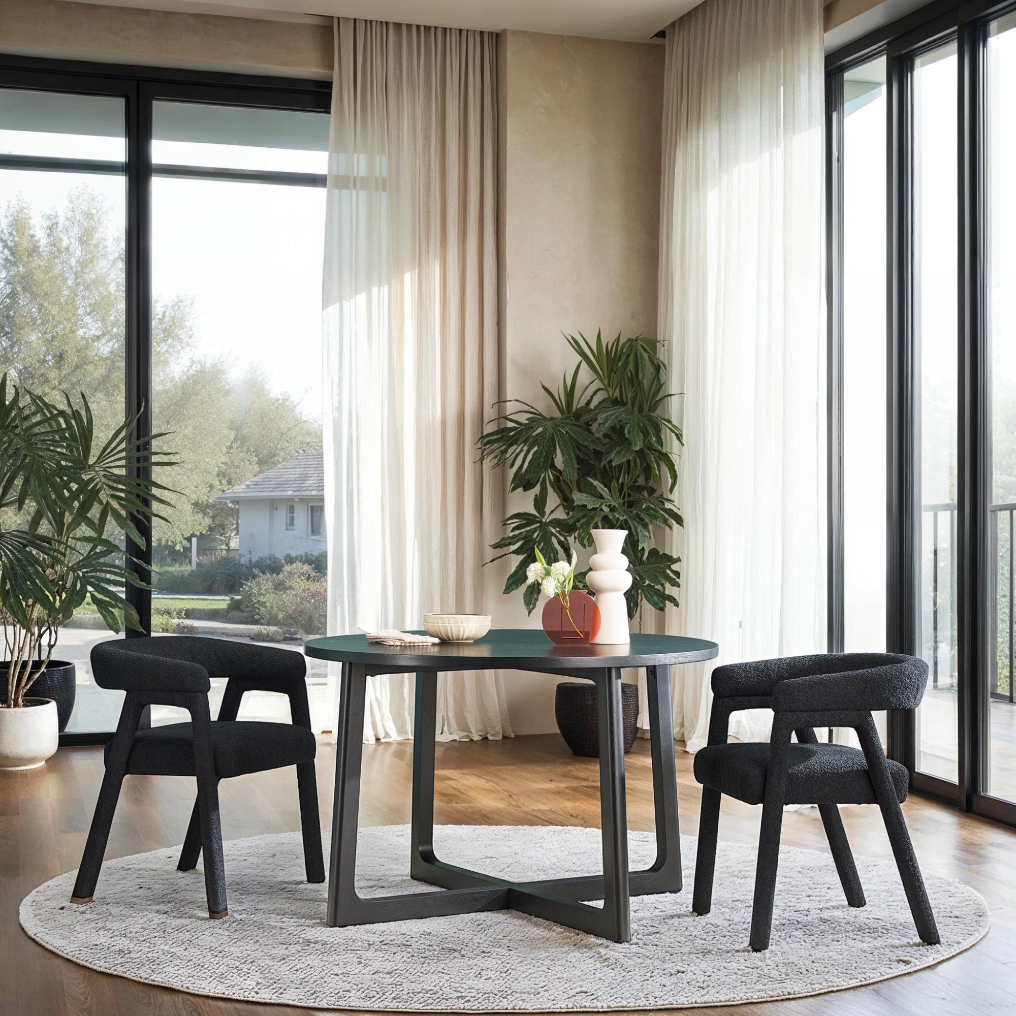 A modern solid wood round dining table with black geometric legs, surrounded by four chairs with dark fabric upholstery. The table is set in a room with large windows, plants, and a light color scheme.