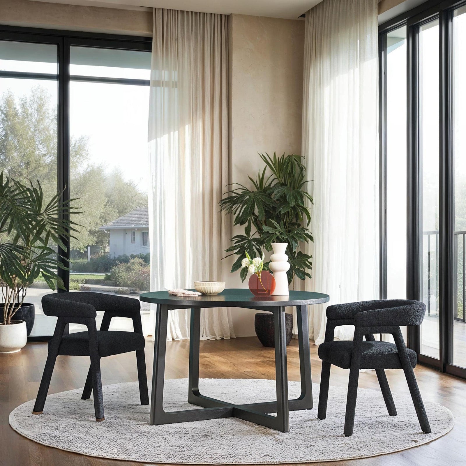 A modern solid wood round dining table with black geometric legs, surrounded by four chairs with dark fabric upholstery. The table is set in a room with large windows, plants, and a light color scheme.