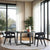 A modern solid wood round dining table with black geometric legs, surrounded by four chairs with dark fabric upholstery. The table is set in a room with large windows, plants, and a light color scheme.
