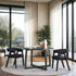 A modern solid wood round dining table with black geometric legs, surrounded by four chairs with dark fabric upholstery. The table is set in a room with large windows, plants, and a light color scheme.